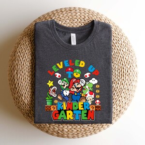以下が含まれることがあります： スーパーマリオブラザーズのビデオゲームシリーズのキャラクター、マリオとその仲間たちのグラフィックが描かれたダークグレーのTシャツ。グラフィックには「LEVELED UP TO KINDERGARTEN」というテキストが含まれています。