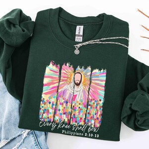 May include: Dark green sweatshirt with a colorful graphic of a religious figure, surrounded by a radiant halo and a crowd of people. The text "Every knee shall bow" and "Philippians 2:10-13" are printed below the image. A silver necklace with a pendant is draped across the sweatshirt.