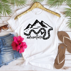K&ouml;nnte beinhalten: Wei&szlig;es T-Shirt mit einer schwarzen Grafik von Bergen und einer kurvigen Stra&szlig;e mit dem Wort "Abenteuer" darunter. Die Grafik hat eine glitzernde Textur.