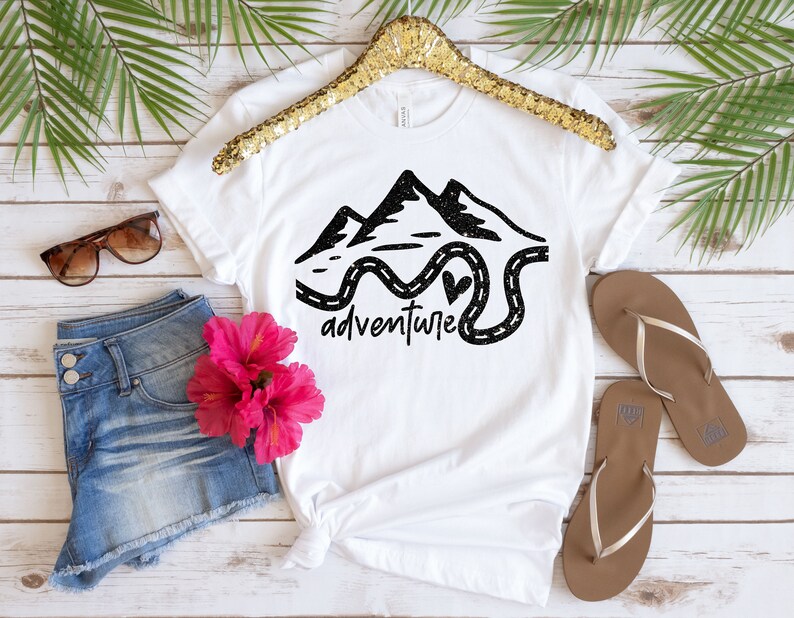 K&ouml;nnte beinhalten: Wei&szlig;es T-Shirt mit einer schwarzen Grafik von Bergen und einer kurvigen Stra&szlig;e mit dem Wort "Abenteuer" darunter. Die Grafik hat eine glitzernde Textur.