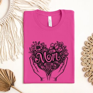Op de afbeelding: Roze T-shirt met een zwarte afbeelding van een hart met het woord "Mom" erin. Het hart is omringd door bloemen en bladeren en wordt vastgehouden door twee handen.