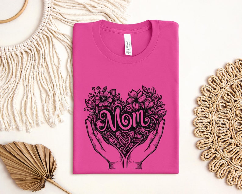 Op de afbeelding: Roze T-shirt met een zwarte afbeelding van een hart met het woord "Mom" erin. Het hart is omringd door bloemen en bladeren en wordt vastgehouden door twee handen.
