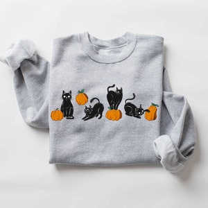 Puede incluir: Una sudadera gris con un dise&ntilde;o de gato negro y calabaza naranja. Los gatos est&aacute;n en varias poses, incluyendo sentados, de pie y acostados. Las calabazas son todas naranjas y tienen tallos verdes.
