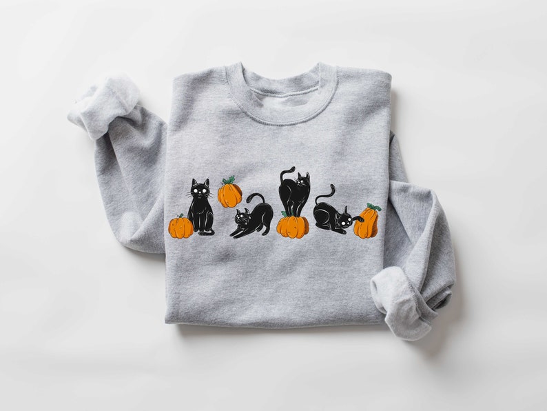 Puede incluir: Una sudadera gris con un dise&ntilde;o de gato negro y calabaza naranja. Los gatos est&aacute;n en varias poses, incluyendo sentados, de pie y acostados. Las calabazas son todas naranjas y tienen tallos verdes.