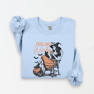 May include: Light blue sweatshirt with a graphic of a witch sitting in a chair with a black cat, pumpkin, and bats. The text "Feeling Witchy" is in orange script. The witch is wearing a black hat and orange dress.