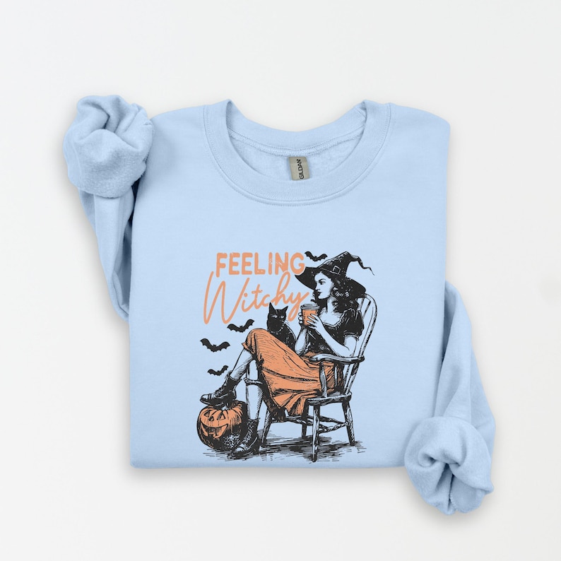 May include: Light blue sweatshirt with a graphic of a witch sitting in a chair with a black cat, pumpkin, and bats. The text "Feeling Witchy" is in orange script. The witch is wearing a black hat and orange dress.