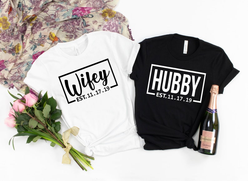 May include: Two white t-shirts with black text. The left shirt says "Wifey - EST. 11.17.19" and the right shirt says "Hubby - EST. 11.17.19".