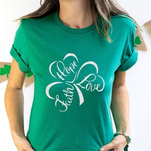 Puede incluir: Una camiseta verde con un dise&ntilde;o de tr&eacute;bol blanco. El tr&eacute;bol tiene las palabras "Fe", "Esperanza" y "Amor" escritas en su interior.
