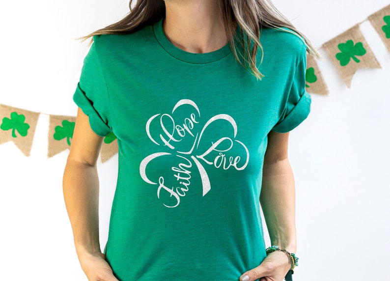 Puede incluir: Una camiseta verde con un dise&ntilde;o de tr&eacute;bol blanco. El tr&eacute;bol tiene las palabras "Fe", "Esperanza" y "Amor" escritas en su interior.