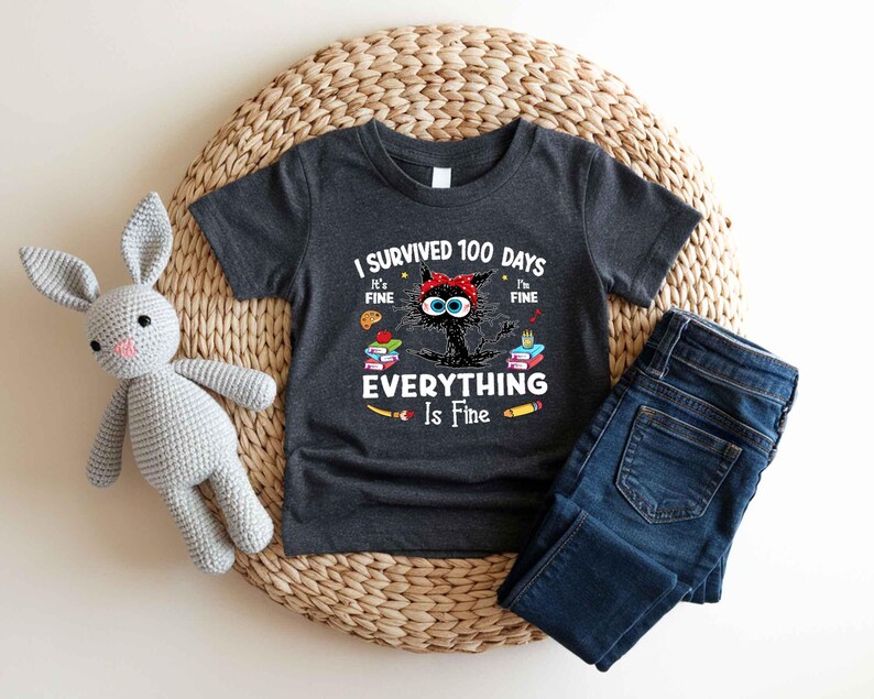 May include: Dark gray t-shirt with the text "I Survived 100 Days Everything Is Fine" and a cartoon cat wearing a red bow. The shirt is paired with blue jeans and rests on a woven mat with a gray stuffed bunny.