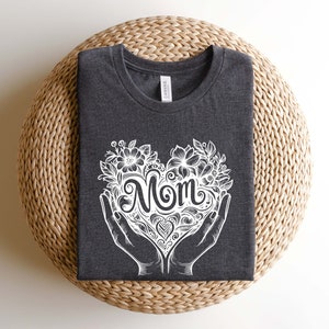 Op de afbeelding: Een donkergrijs T-shirt met een wit ontwerp van een hart met het woord "Mom" erin. Het hart is omringd door bloemen en bladeren.