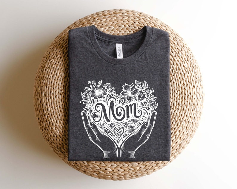 Op de afbeelding: Een donkergrijs T-shirt met een wit ontwerp van een hart met het woord "Mom" erin. Het hart is omringd door bloemen en bladeren.