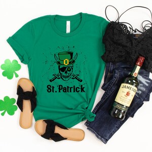 May include: Green t-shirt with a black and white skull wearing a green leprechaun hat and holding crossed swords. The text "St. Patrick" is below the skull. The shirt is laid out on a white surface with a pair of black sandals, a black lace bralette, blue jeans, and a bottle of Jameson Irish Whiskey.