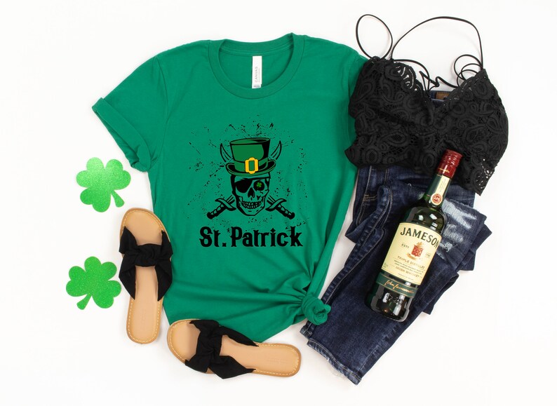 May include: Green t-shirt with a black and white skull wearing a green leprechaun hat and holding crossed swords. The text "St. Patrick" is below the skull. The shirt is laid out on a white surface with a pair of black sandals, a black lace bralette, blue jeans, and a bottle of Jameson Irish Whiskey.