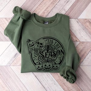 May include: A green sweatshirt with a black graphic print of a skeleton holding a pumpkin and the text "Tis the Season".