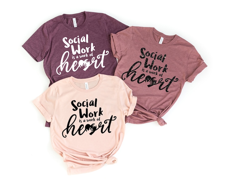 May include: Three t-shirts with the text "Social Work is a work of heart" in black lettering with a handprint heart. The shirts are in different shades of pink and purple.