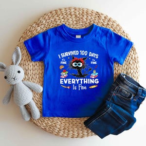 May include: A royal blue t-shirt with the text "I Survived 100 Days, It's Fine, I'm Fine, Everything Is Fine" and a cartoon cat design. The cat has a red bow and is surrounded by books and school supplies. Dark blue jeans are folded next to the shirt.