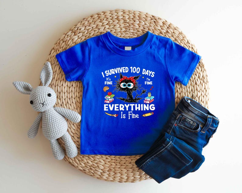 May include: A royal blue t-shirt with the text "I Survived 100 Days, It's Fine, I'm Fine, Everything Is Fine" and a cartoon cat design. The cat has a red bow and is surrounded by books and school supplies. Dark blue jeans are folded next to the shirt.