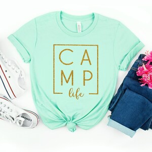 May include: A light green t-shirt with a gold glitter graphic that says "CAMP life" in a box.