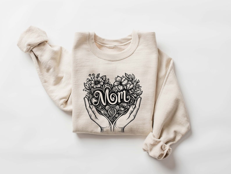 Op de afbeelding: Een beige sweatshirt met een zwarte afbeelding van een hart met het woord "MOM" erin, omringd door bloemen en vastgehouden door twee handen.