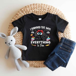 May include: Black t-shirt with the text "I Survived 100 Days Everything Is Fine" and a cartoon cat wearing a red headband. The shirt is paired with blue jeans and a gray stuffed bunny.