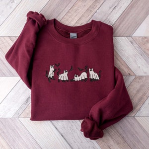 Pode incluir: Um moletom bord&ocirc; com um desenho bordado de gatos vestidos de fantasmas. Os gatos est&atilde;o em v&aacute;rias poses, com morcegos voando acima deles. O moletom tem gola redonda e mangas compridas.
