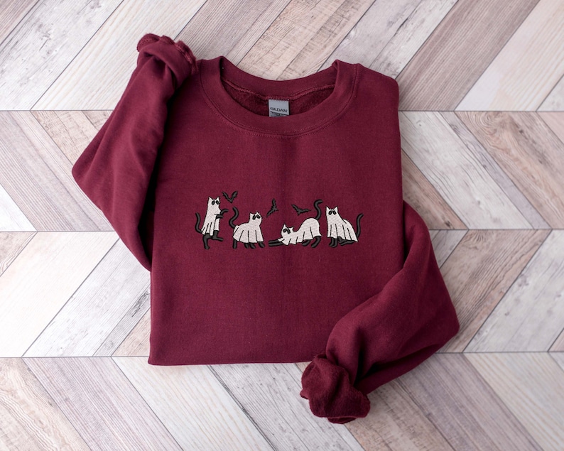 Pode incluir: Um moletom bord&ocirc; com um desenho bordado de gatos vestidos de fantasmas. Os gatos est&atilde;o em v&aacute;rias poses, com morcegos voando acima deles. O moletom tem gola redonda e mangas compridas.