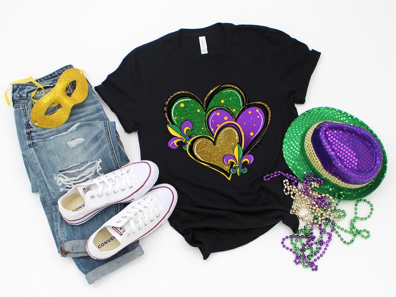 May include: A black t-shirt featuring a gold, green and purple heart design. The design incorporates two hearts with a fleur-de-lis. The shirt is laid flat on a white surface with a pair of white trainers, a pair of blue jeans, a gold mask, a green and purple hat, and a string of beads.