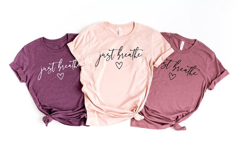 Puede incluir: Tres camisetas con el texto "just breathe" y un peque&ntilde;o dise&ntilde;o de coraz&oacute;n. Las camisetas son en tonos de rosa y morado.