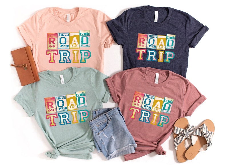 May include: Four t-shirts with a vintage road trip design. The shirts are in different colors: pink, blue, green, and brown. The design features a colorful graphic of the word "ROAD" with a vintage license plate design and the word "TRIP" underneath.