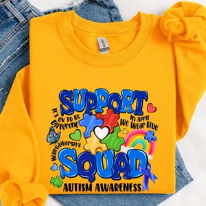 May include: Mustard yellow sweatshirt with the words "SUPPORT SQUAD AUTISM AWARENESS" in blue, white, and red. The design includes puzzle pieces, a rainbow, a butterfly, and the phrase "In April We Wear Blue".