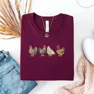May include: Burgundy crew neck t-shirt featuring an embroidered design of four chickens in a row. The shirt is paired with blue jeans, white slip-on shoes, and a necklace. A dried palm leaf is also in the image.
