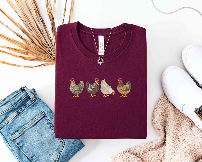May include: Burgundy crew neck t-shirt featuring an embroidered design of four chickens in a row. The shirt is paired with blue jeans, white slip-on shoes, and a necklace. A dried palm leaf is also in the image.