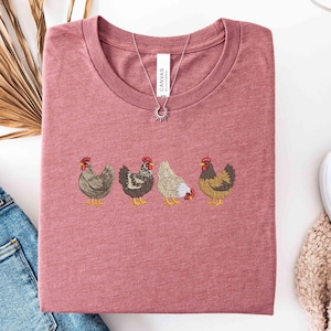 May include: A dusty rose t-shirt with an embroidered design of four chickens in various colors. The shirt has a crew neck and is folded. A silver necklace with a sun pendant is on top of the shirt.