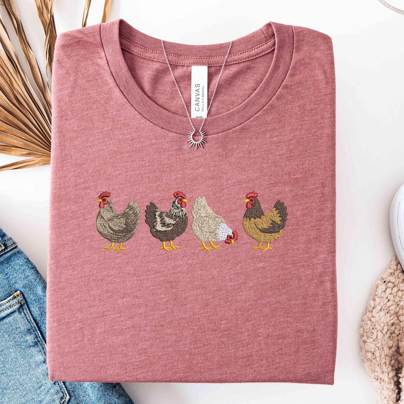 May include: A dusty rose t-shirt with an embroidered design of four chickens in various colors. The shirt has a crew neck and is folded. A silver necklace with a sun pendant is on top of the shirt.