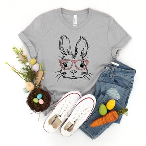 May include: A gray t-shirt with a black and white illustration of a bunny wearing pink glasses. The shirt is paired with blue jeans, white sneakers, and a nest with colorful Easter eggs.