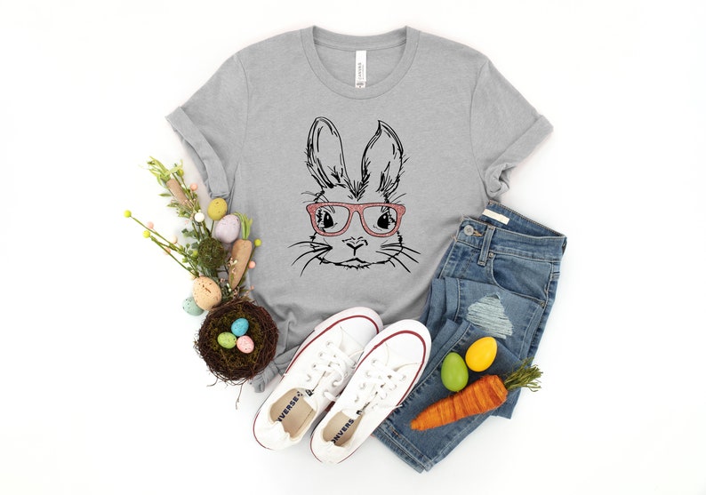 May include: A gray t-shirt with a black and white illustration of a bunny wearing pink glasses. The shirt is paired with blue jeans, white sneakers, and a nest with colorful Easter eggs.