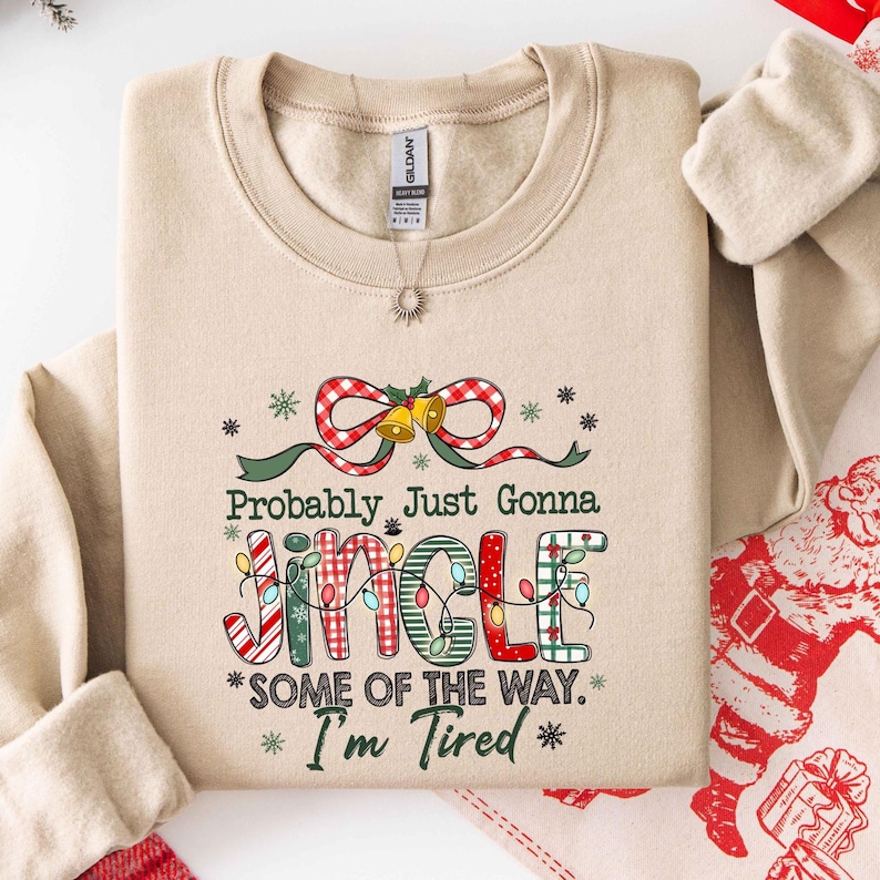 Pu&ograve; includere: Felpa beige con la scritta "Probably Just Gonna Jingle Some of the Way. I'm Tired". La parola "Jingle" &egrave; decorata con luci e motivi natalizi. Un fiocco a scacchi rosso e bianco e delle campane sono in cima.