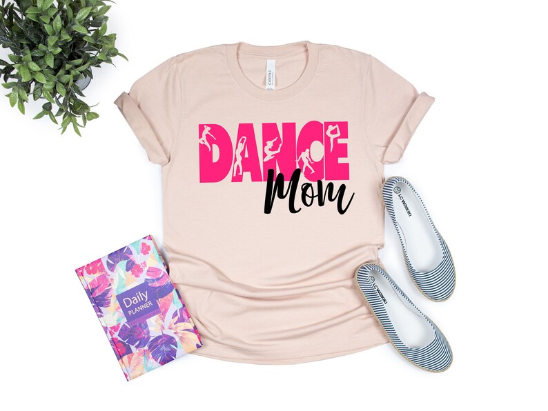 May include: A light pink t-shirt with the text "Dance Mom" in black and pink. The word "Dance" is in pink and has silhouettes of dancers in the letters.
