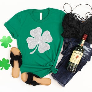 K&ouml;nnte beinhalten: Gr&uuml;nes T-Shirt mit einem vierbl&auml;ttrigen Kleeblatt aus silbernem Glitzer. Das Shirt wird mit einer blauen Jeans, einem schwarzen Spitzen-BH und schwarzen Sandalen kombiniert. Eine Flasche Jameson Irish Whiskey ist ebenfalls im Bild zu sehen.