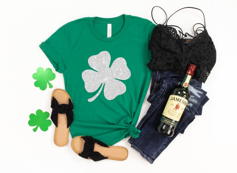 K&ouml;nnte beinhalten: Gr&uuml;nes T-Shirt mit einem vierbl&auml;ttrigen Kleeblatt aus silbernem Glitzer. Das Shirt wird mit einer blauen Jeans, einem schwarzen Spitzen-BH und schwarzen Sandalen kombiniert. Eine Flasche Jameson Irish Whiskey ist ebenfalls im Bild zu sehen.