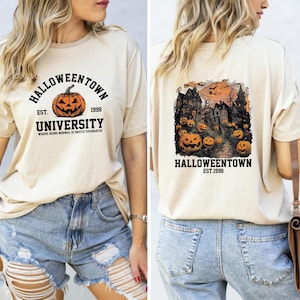 May include: A beige t-shirt with a graphic print of a pumpkin and the text "Halloweentown University Est. 1998 Where being normal is vastly overrated". The back of the shirt has a graphic print of a spooky town with houses and pumpkins.