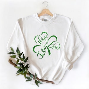 Puede incluir: Sudadera blanca con un dise&ntilde;o de tr&eacute;bol verde que dice "Faith Hope Love" en una fuente cursiva.