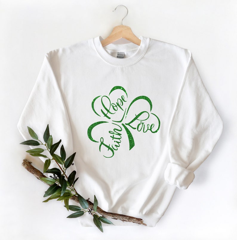 Puede incluir: Sudadera blanca con un dise&ntilde;o de tr&eacute;bol verde que dice "Faith Hope Love" en una fuente cursiva.