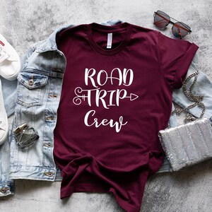 May include: A maroon t-shirt with white text that reads "Road Trip Crew" with a decorative arrow. The shirt is laid flat on a gray textured surface with a denim jacket, white sneakers, sunglasses, and a silver sequined purse.