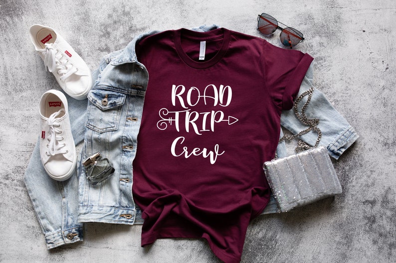 May include: A maroon t-shirt with white text that reads "Road Trip Crew" with a decorative arrow. The shirt is laid flat on a gray textured surface with a denim jacket, white sneakers, sunglasses, and a silver sequined purse.