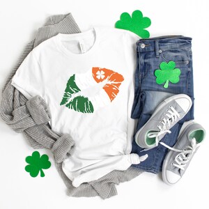 May include: A white t-shirt with a green, white, and orange lip print, resembling the Irish flag, is displayed with blue jeans, gray sneakers, and a gray sweater. Green shamrock decorations are also present, suggesting a St. Patrick's Day theme.