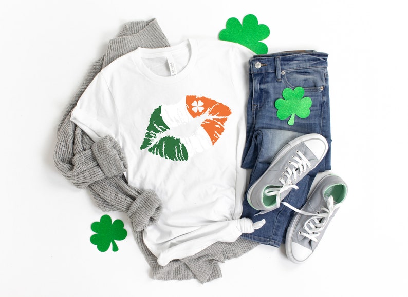 May include: A white t-shirt with a green, white, and orange lip print, resembling the Irish flag, is displayed with blue jeans, gray sneakers, and a gray sweater. Green shamrock decorations are also present, suggesting a St. Patrick's Day theme.
