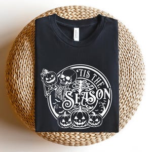 May include: Black t-shirt with a white graphic design of a skeleton holding a pumpkin and the text "Tis the Season".