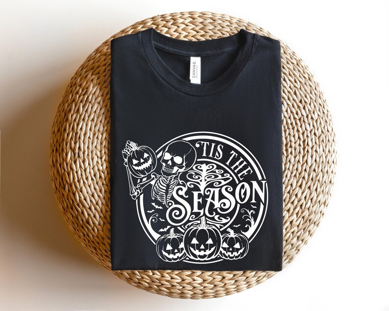 May include: Black t-shirt with a white graphic design of a skeleton holding a pumpkin and the text "Tis the Season".